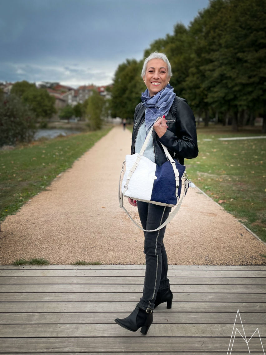 Photo d'une femme blanche mûre portant un sac d'épaule bleu marine et blanc, dans un parc en extérieur, de jour.
