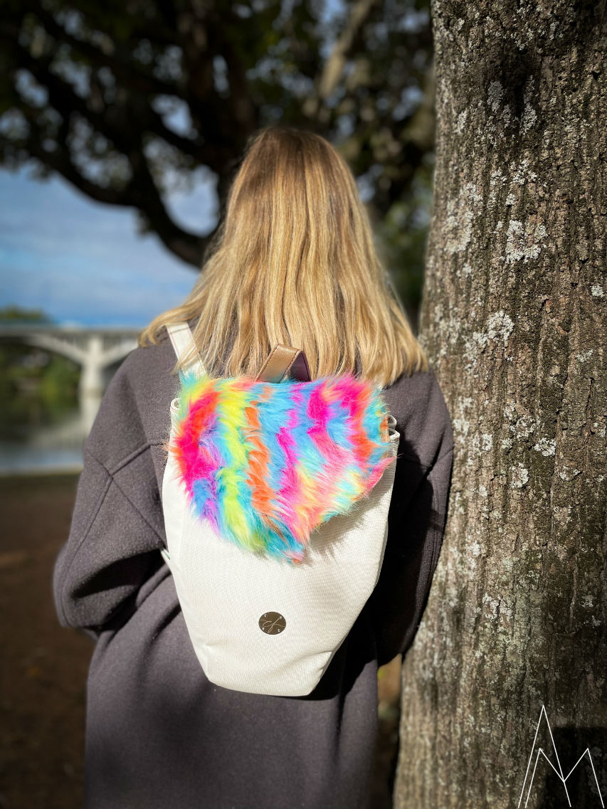Photo d'une jeune femme blonde vue de dos, portant un sac à dos blanc et arc en ciel, dans un parc en extérieur, de jour.