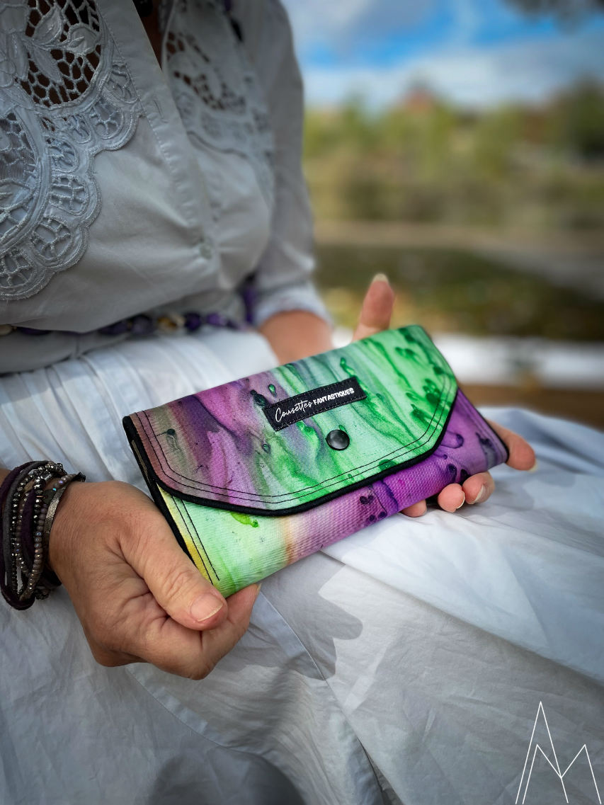 Photo d'une femme blanche mûre tenant un portefeuille dans les tons verts et violets, dans un parc en extérieur, de jour.