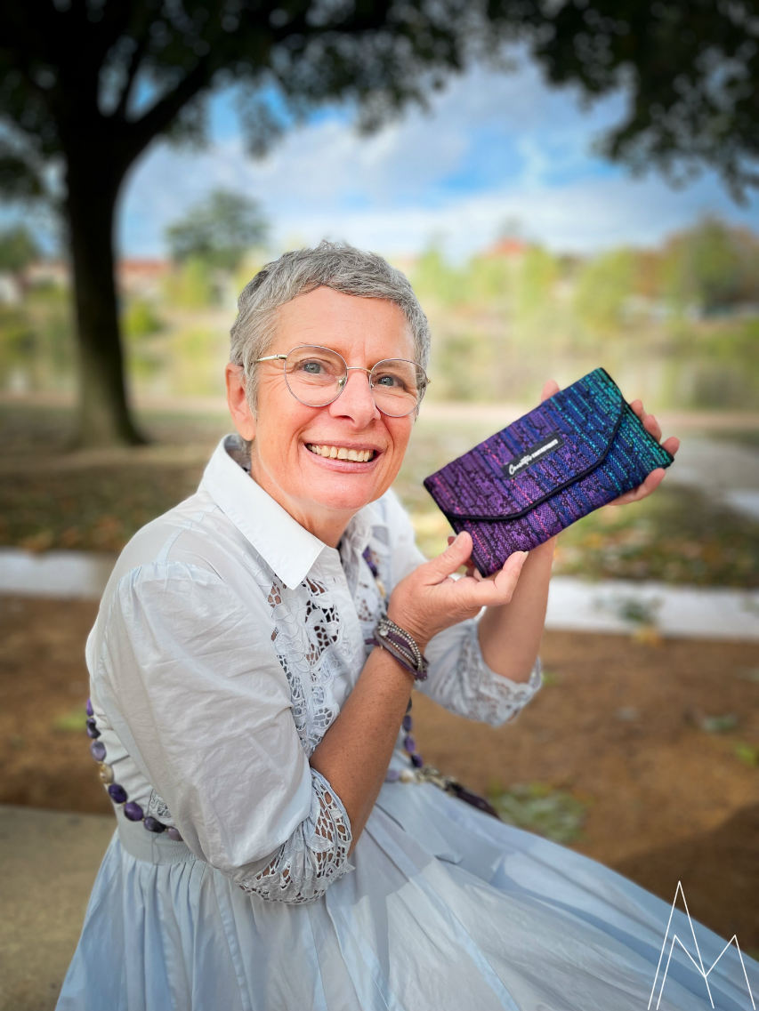 Photo d'une femme blanche mûre portant un portefeuille dans les tons violet et bleus, dans un parc en extérieur, de jour.