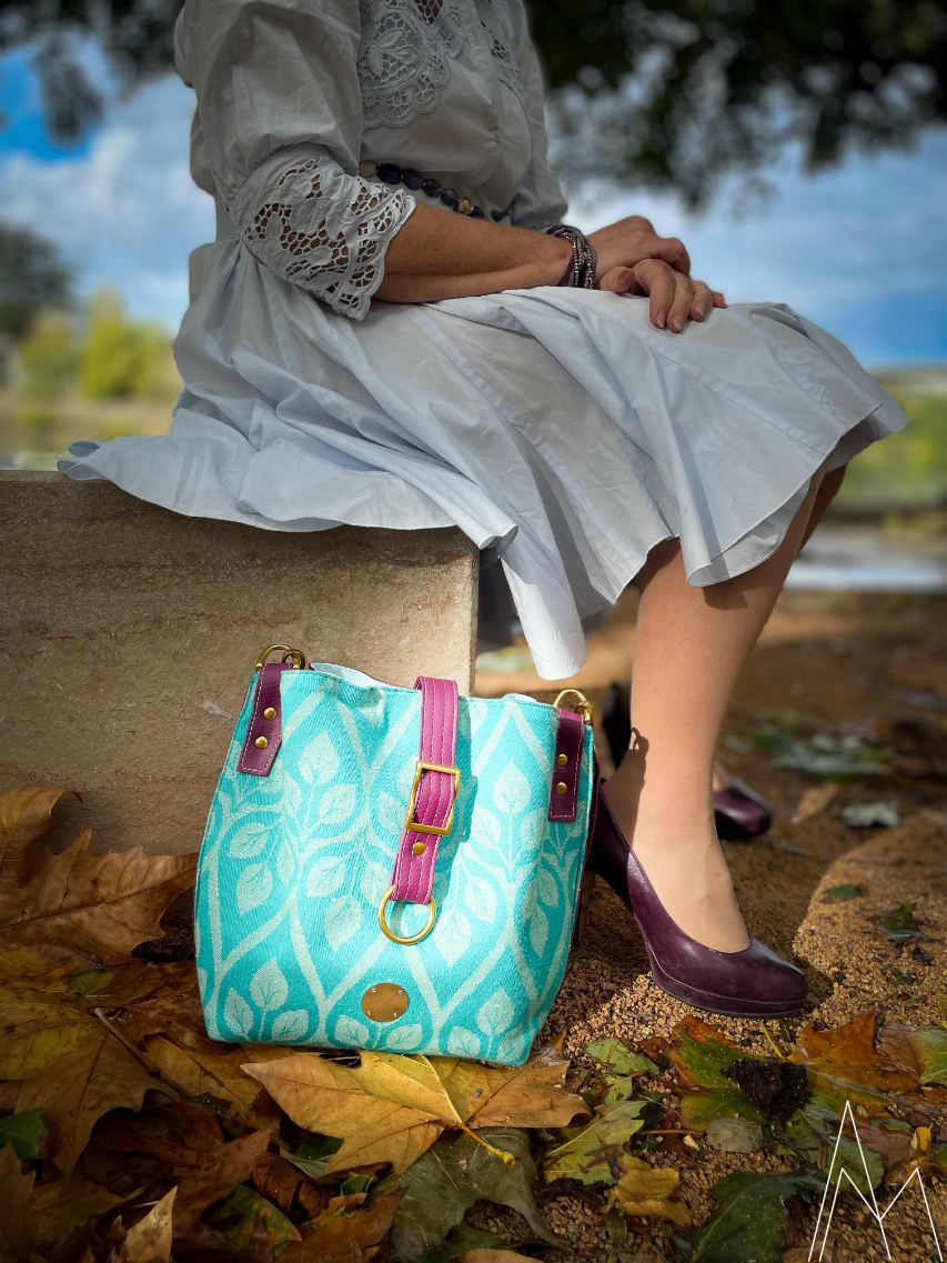 Photo d'une femme blanche mûre assise avec à ses pieds un sac d'épaule turquoise et prune, dans un parc en extérieur, de jour.