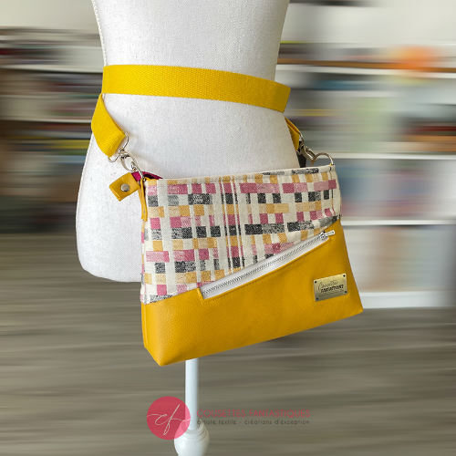 A rectangular messenger bag made from black, white, pink, and yellow babywearing wrap fabric with a geometric pattern, and sunflower yellow faux leather.
