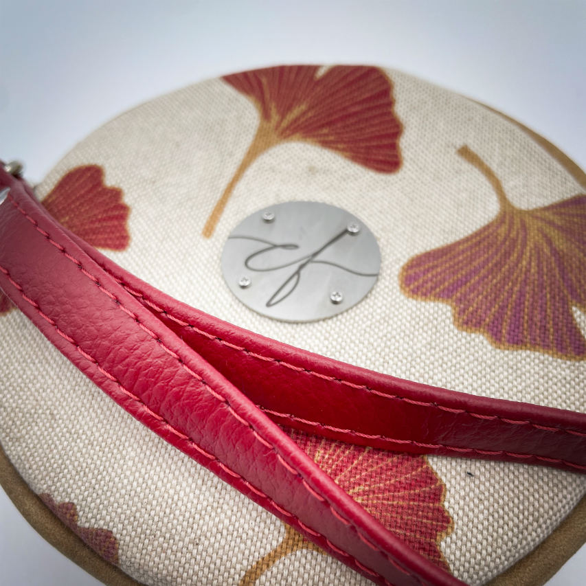 Une pochette ronde faite de cuir de couleur camel et d'une toile en polycoton avec un motif de feuilles de Ginkgo Biloba rouges.