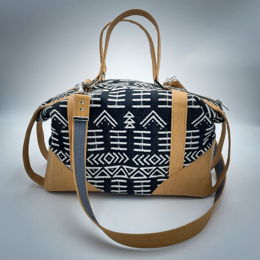 Un petit sac de voyage fabriqué en papier lavable couleur caramel, une écharpe de portage à motif géométrique noir et blanc, et de la popeline grise.