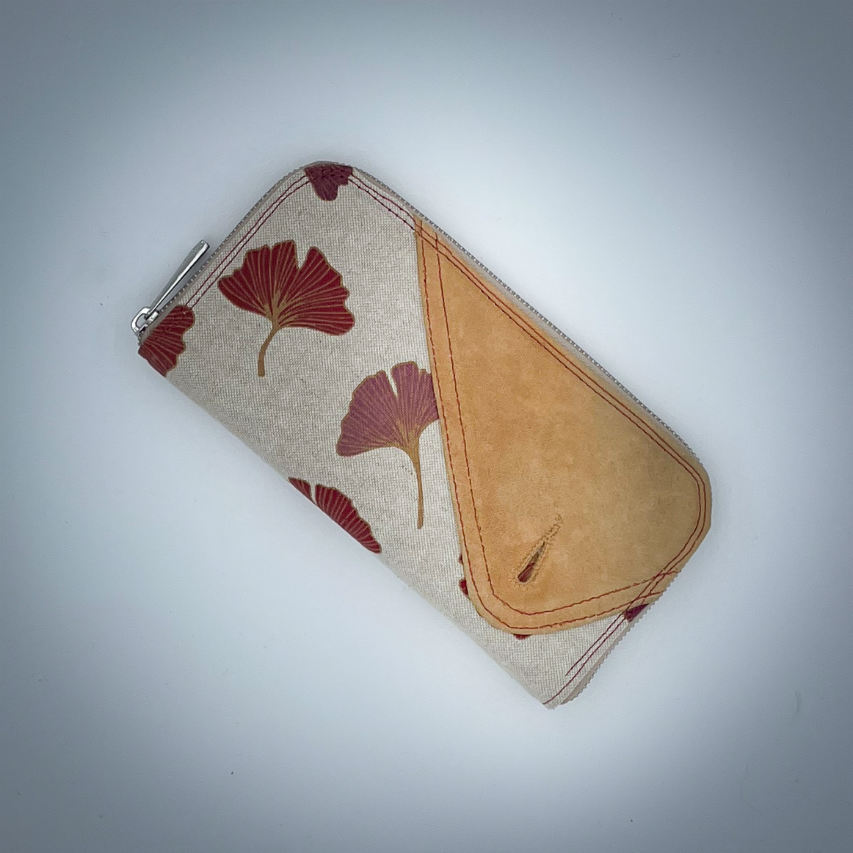Un portefeuille zippé fait de cuir de couleur camel et d'une toile en polycoton avec un motif de feuilles de Ginkgo Biloba rouges.