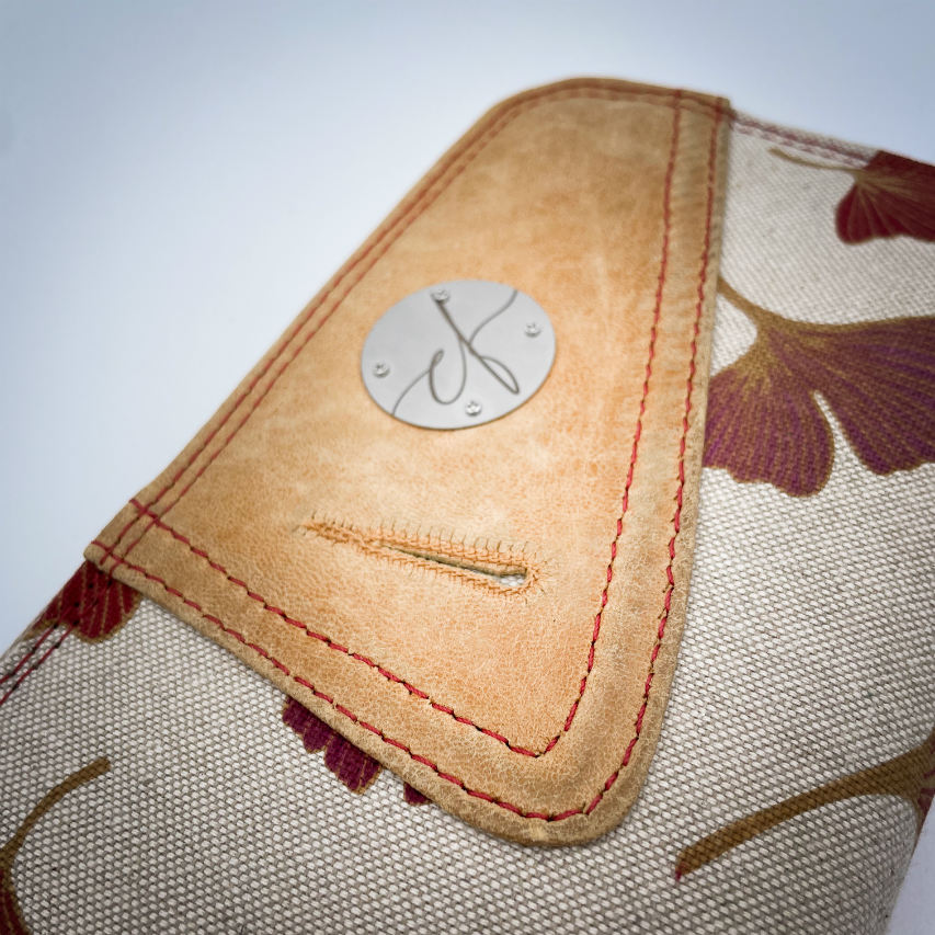 Un portefeuille zippé fait de cuir de couleur camel et d'une toile en polycoton avec un motif de feuilles de Ginkgo Biloba rouges.