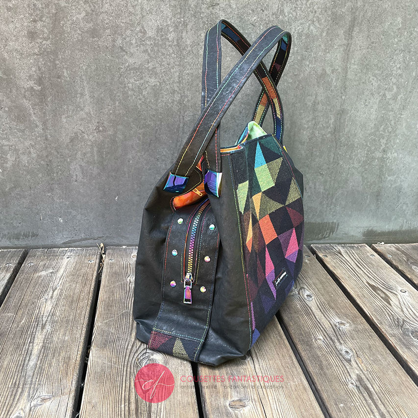 A shoulder bag made from black and rainbow geometric-patterned babywearing scarf and black cork, with a bright rainbow swirl interior.