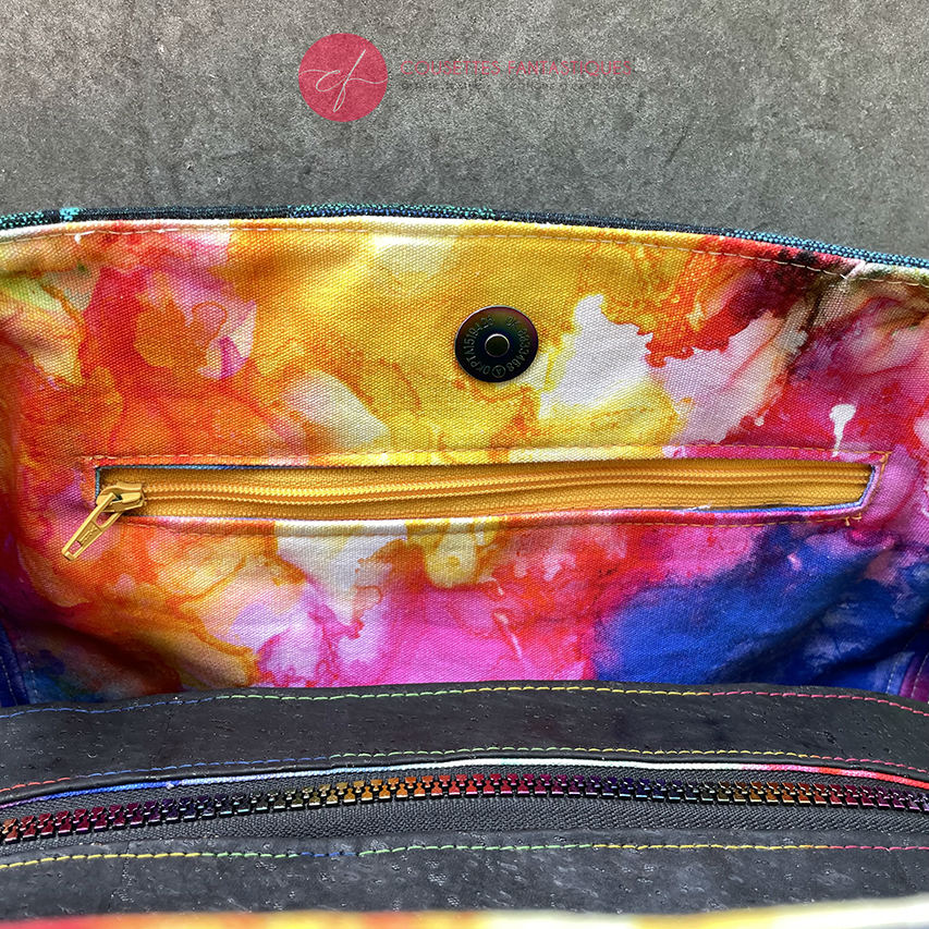 A shoulder bag made from black and rainbow geometric-patterned babywearing scarf and black cork, with a bright rainbow swirl interior.