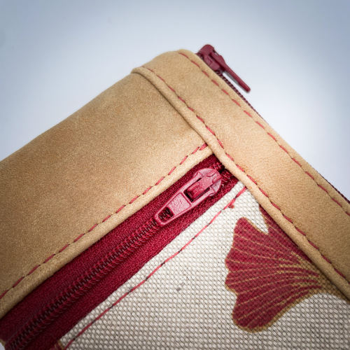 Un mini portemonnaie zippé fait de cuir de couleur camel et d'une toile en polycoton avec un motif de feuilles de Ginkgo Biloba rouges.